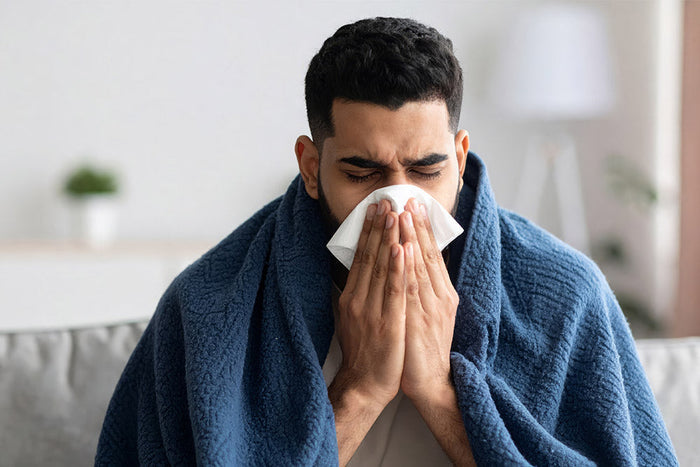 A man wrapped in a blue blanket is sitting indoors, holding a tissue to his nose while sneezing or blowing, appearing sick or congested.