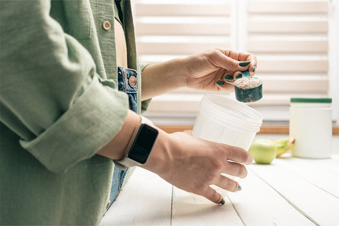 A person scooping protein powder into a shaker bottle, representing HMB supplementation for muscle recovery, strength, and performance support.