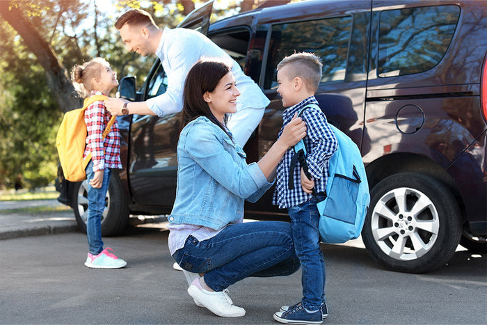 A smiling mother and father help their two young children get ready for school beside a parked car.