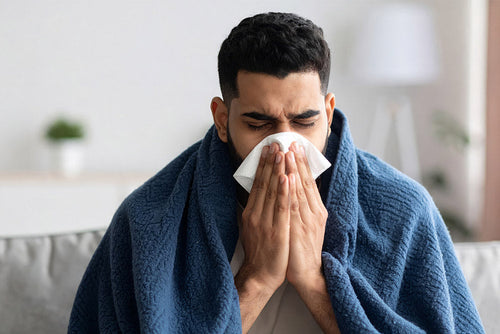 A man wrapped in a blue blanket is sitting indoors, holding a tissue to his nose while sneezing or blowing, appearing sick or congested.