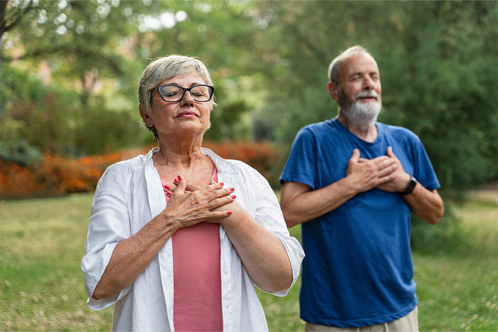 An older woman and man stand outdoors with their eyes closed, hands over their chests, appearing calm and focused while practicing mindfulness or meditation. They are surrounded by greenery and flowers in a peaceful park setting.