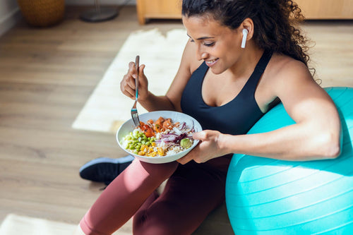 Woman sitting beside an exercise ball and eating a balanced bowl, highlighting the importance of fueling your body to support workouts and recovery.