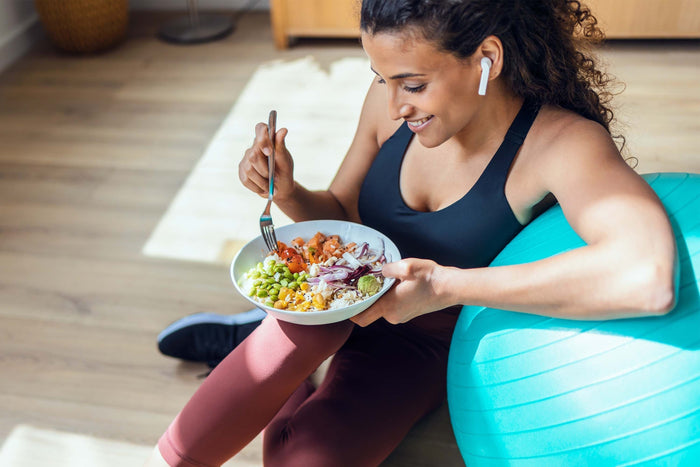 Woman sitting beside an exercise ball and eating a balanced bowl, highlighting the importance of fueling your body to support workouts and recovery.