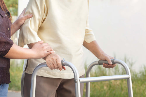 An older adult uses a walker with caregiver assistance, highlighting mobility and nutritional recovery after hip fracture surgery.