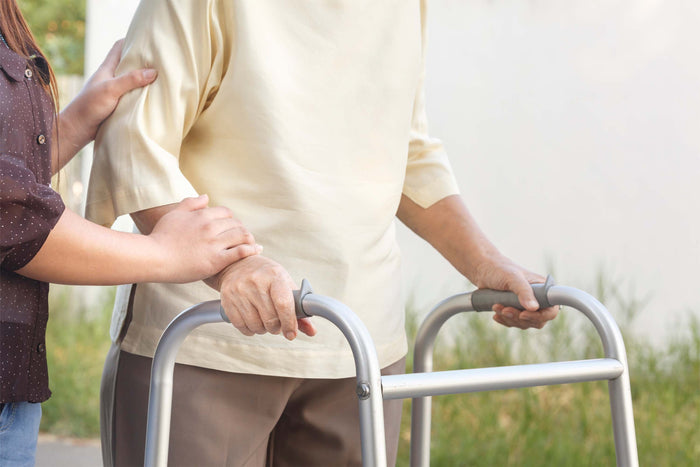 An older adult uses a walker with caregiver assistance, highlighting mobility and nutritional recovery after hip fracture surgery.